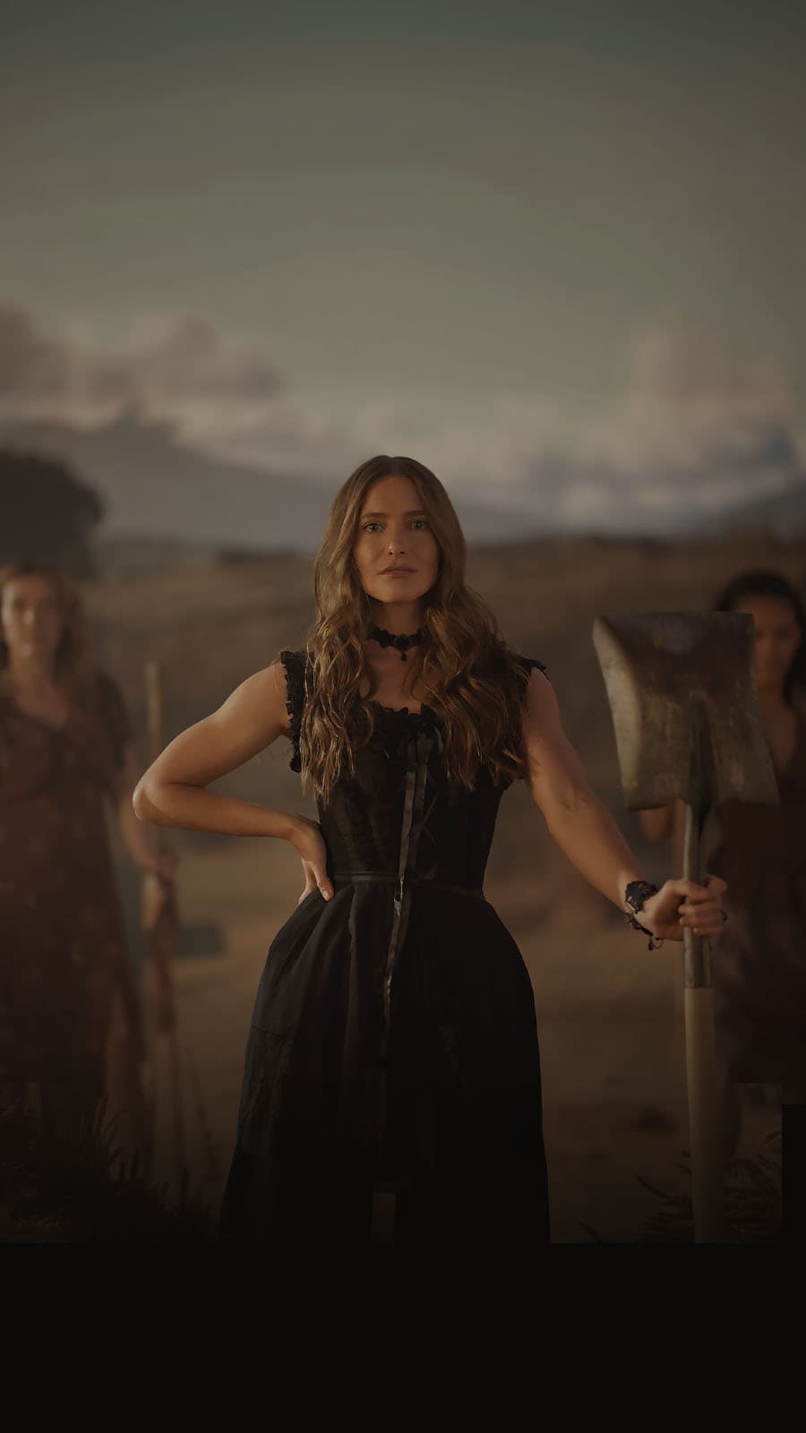 Caroline Jones in a black dress, hand on hip and holding a shovel, in a dusty field with two women behind her.